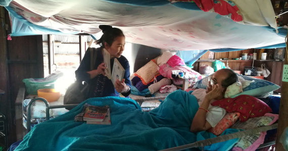 In a room with multiple hospital beds a man is lying in a bed, covered with a blanket and propped up by pillows. He is wearing reading glasses and looking at the librarian who offers different books to read.