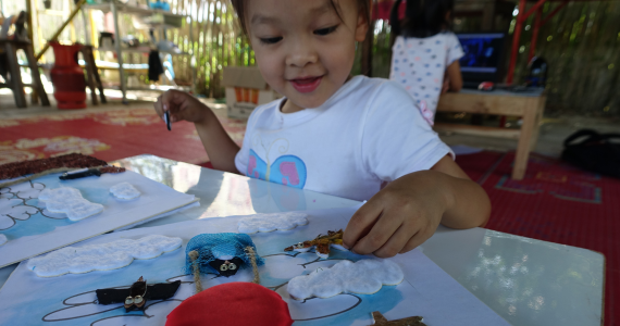 A girl around three years old is sitting at a table and playing with a Noon Noon Book. The pictures on the pages are tactile and interactive. Clouds are made out of cotton and a penguin puppet is sitting in a hot air balloon made out of mesh. 