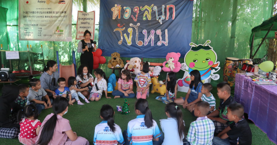 20 children sit in a circle outside in front of the library. They are looking at a librarian who is shaking two rattles. In the background are toys, a trampoline, a slide and a table with craft materials.